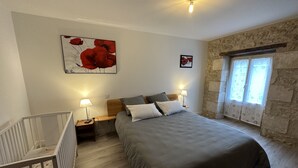 A bedroom with a large bed, two bedside tables with lamps, a crib, a stone wall, and a window with curtains.