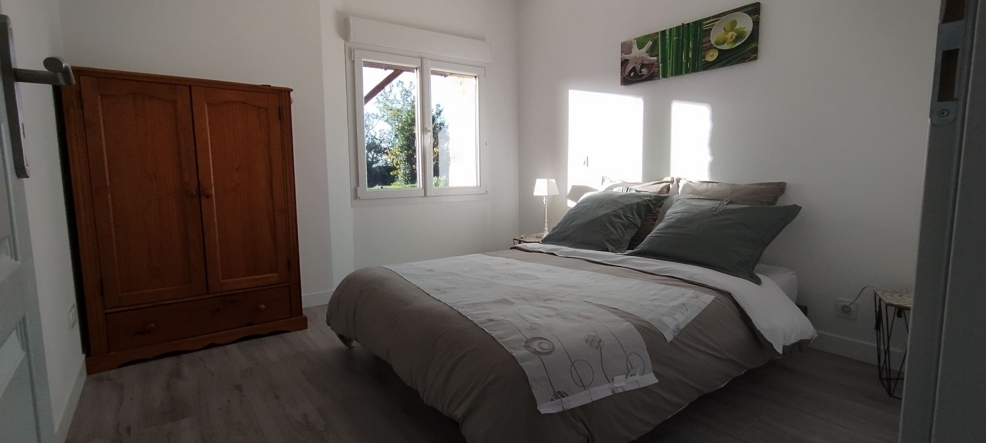 A bedroom with a bed, a wooden wardrobe, a window, and a wall-mounted picture.