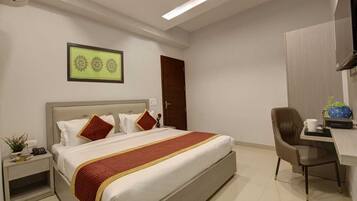 A hotel room with a bed, a desk, a chair, a small table, a wall-mounted TV, and a door.