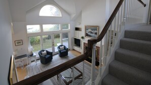 A spacious living area with a high ceiling, a fireplace, and a staircase leading to an upper floor. - Pet Friendly Spacious Waterfront Home With A Deep Water Access Dock (2324) (Cotuit)