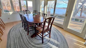 A dining room with a wooden table and chairs, large windows, and a view of a body of water. - Pet Friendly Spacious Waterfront Home With A Deep Water Access Dock (2324) (Cotuit)