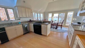 A spacious kitchen with wooden floors, white cabinets, a built-in dishwasher, and a dining area with a view of the outdoors. - Pet Friendly Spacious Waterfront Home With A Deep Water Access Dock (2324) (Cotuit)