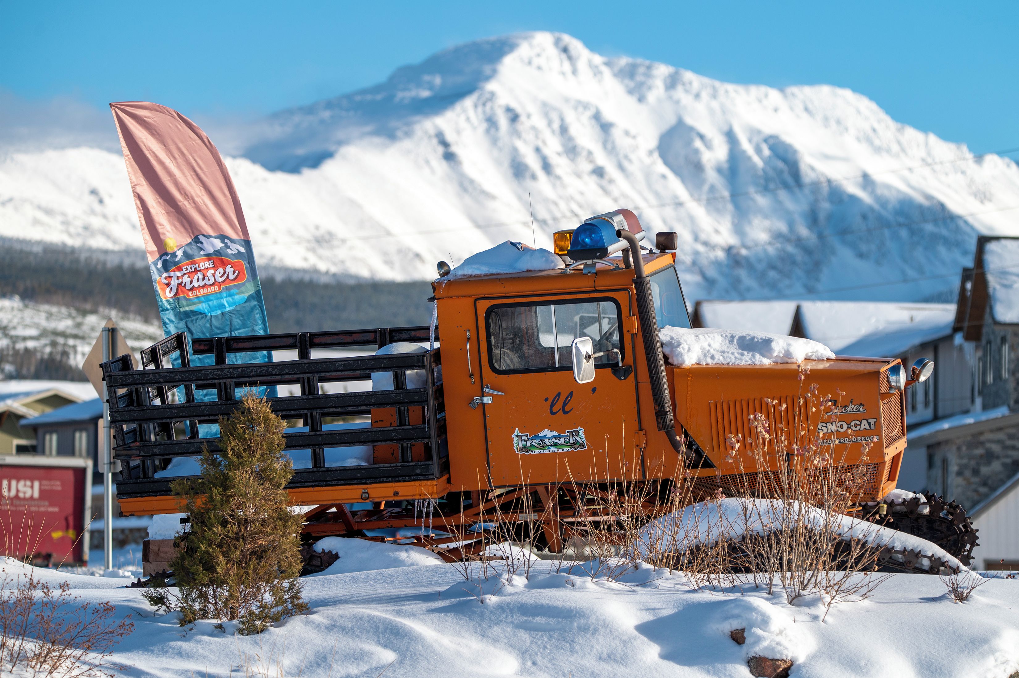 Un camió cobert de neu amb un cartell que diu 'Explore Fraser', estacionat en una zona nevada amb muntanyes al fons.