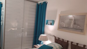 Un dormitorio con cama, mesita de noche, lámpara, un cuadro enmarco y un armario con cortina azul.
