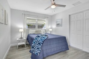 Un dormitorio con una cama, dos mesitas de noche con lámparas, un ventilador de techo, una ventana con cortinas y una puerta.
