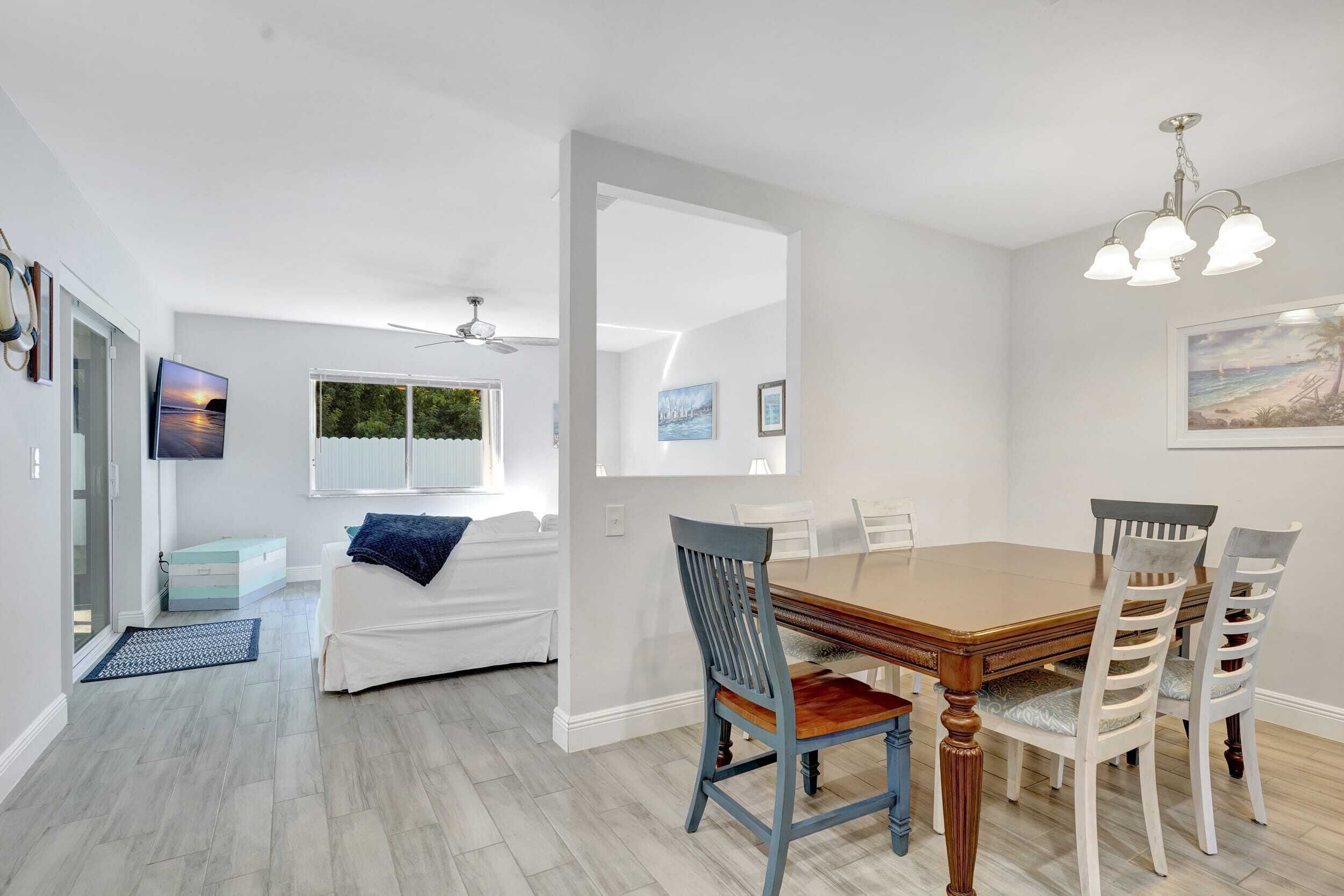 A dining area with a wooden table and white chairs, a ceiling fan, and a view of a bedroom with a bed and a window.