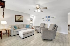 A living room with a sofa, ottoman, two armchairs, a coffee table, a floor lamp, a ceiling fan, and framed pictures on the wall.