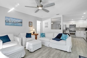 A modern living room with white furniture, a ceiling fan, and a kitchen in the background.
