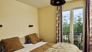 Un dormitorio con cama, dos almohadas, una mesita de noche, ventana con cortinas y balcón con barandilla.
