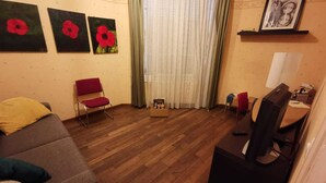 Una sala de estar con un sofá, un sillón rojo, un escritorio con una computadora y una ventana con cortinas.