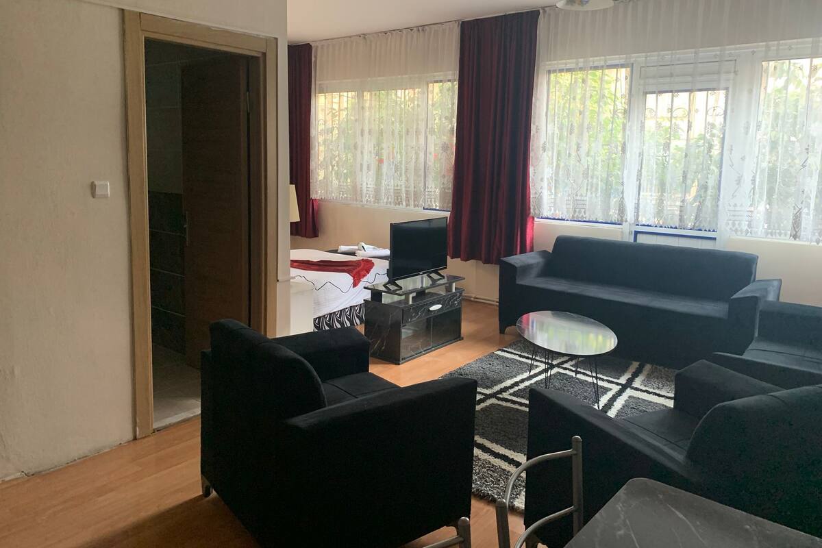 A living room with a sofa, chairs, a TV, and a small table. There is a bed in the background with a window and curtains.