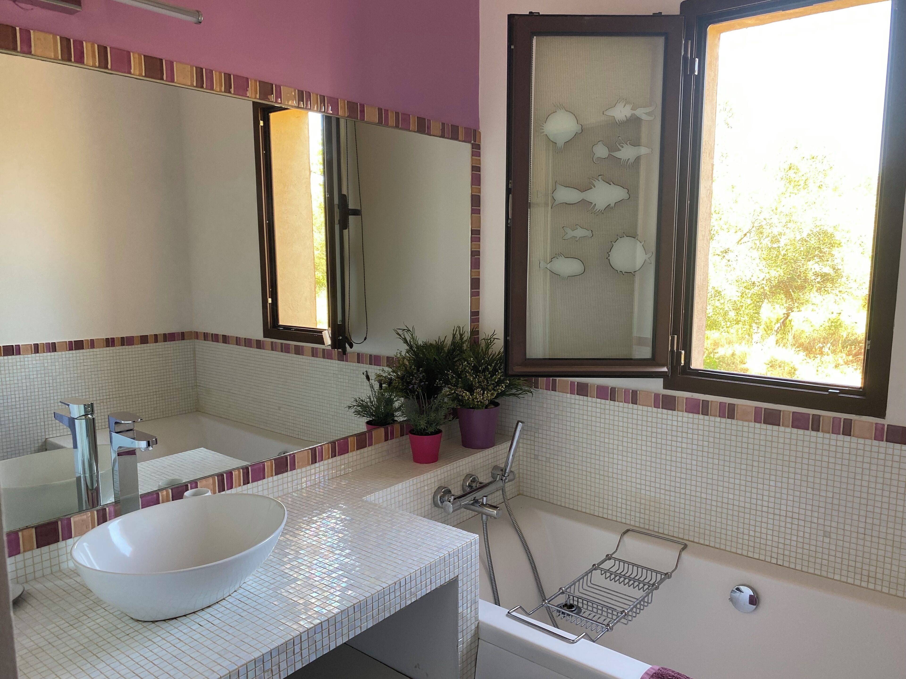 Une salle de bain avec un lavabo blanc, un grand miroir, une fenêtre donnant sur un espace verdoyant et une baignoire équipée d’un pommeau de douche.