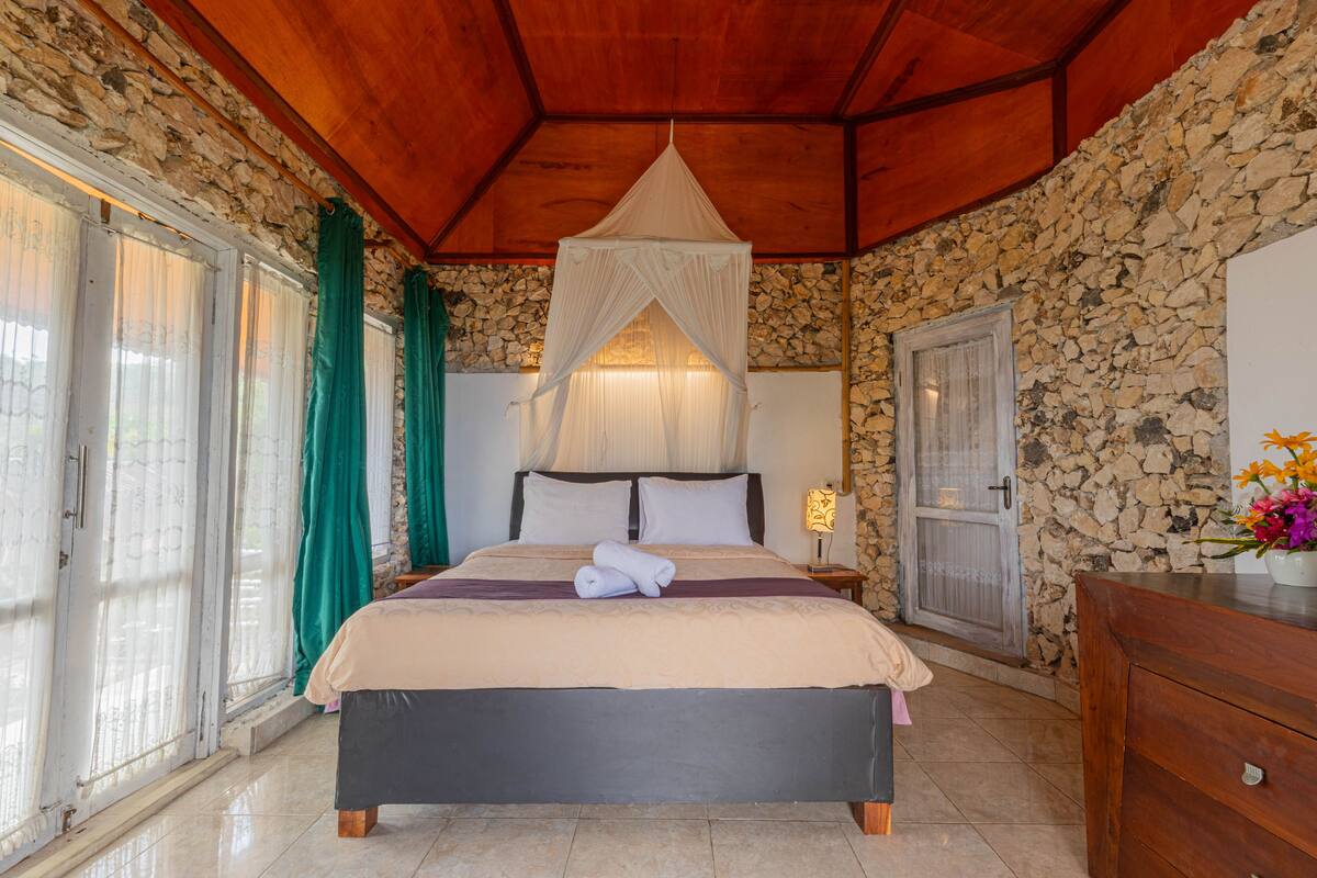 A bedroom with a stone wall, a bed with a canopy, a nightstand, and a dresser.