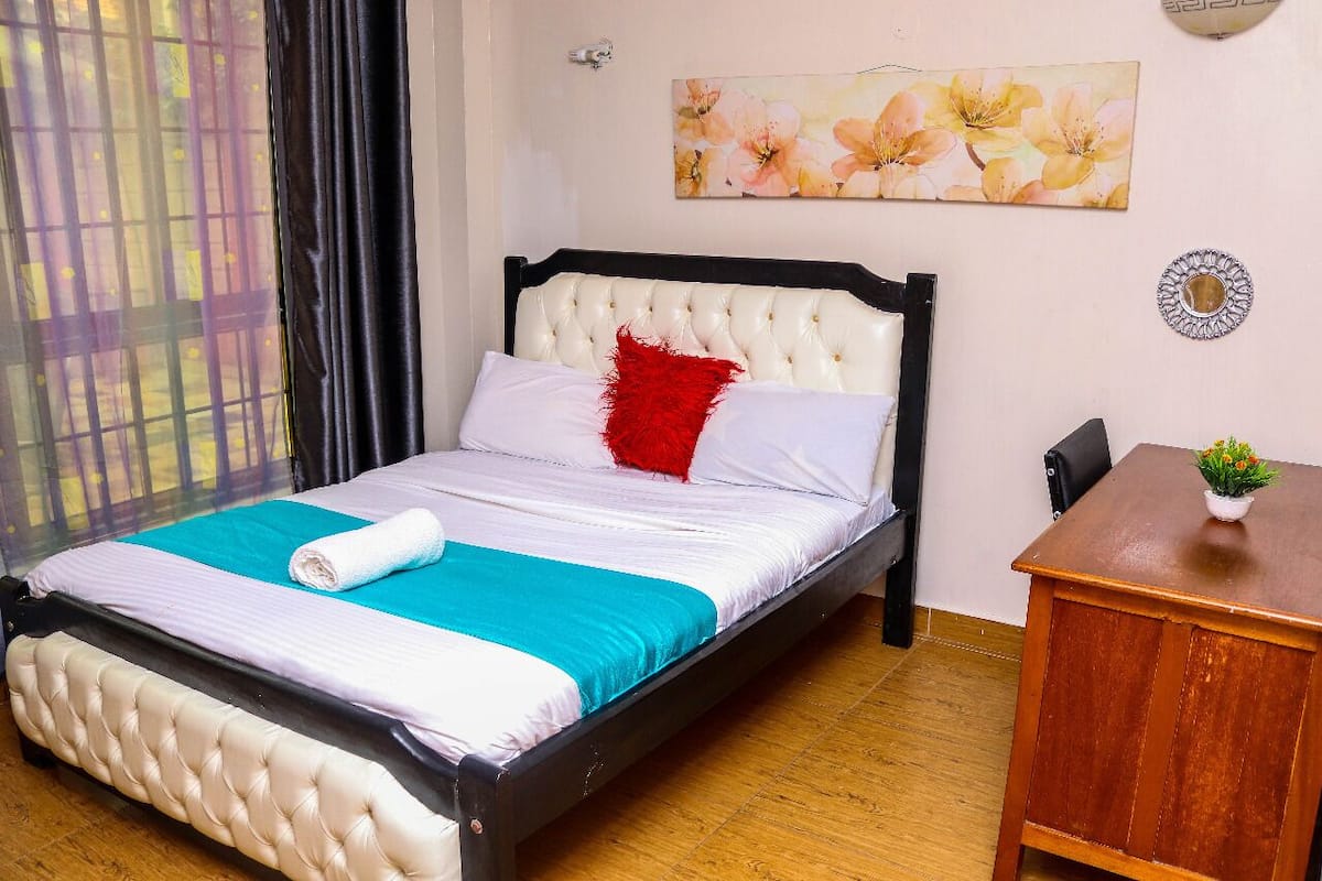 A neatly made bed with a white and teal bedspread, a red pillow, and a rolled white towel. There is a wooden table with a small plant on it, and a chair next to the table.