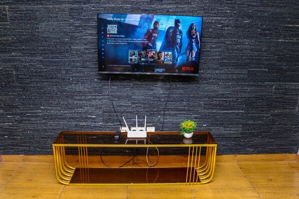 A flat-screen TV mounted on a textured wall displaying a movie selection interface. Below the TV, a wooden console with a plant and a white electronic device is visible.