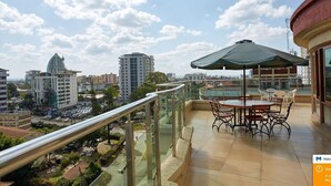 A balcony with a glass railing overlooking a cityscape, featuring a table and chairs, and a large umbrella.
