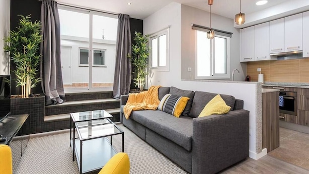Ein modernes Wohnzimmer mit grauem Sofa, gelbem Sessel und einem Glastisch. Zwei große Fenster mit Vorhängen, eine Küchenzeile mit weißen Schränken und ein kleiner Balkon mit Pflanzen.