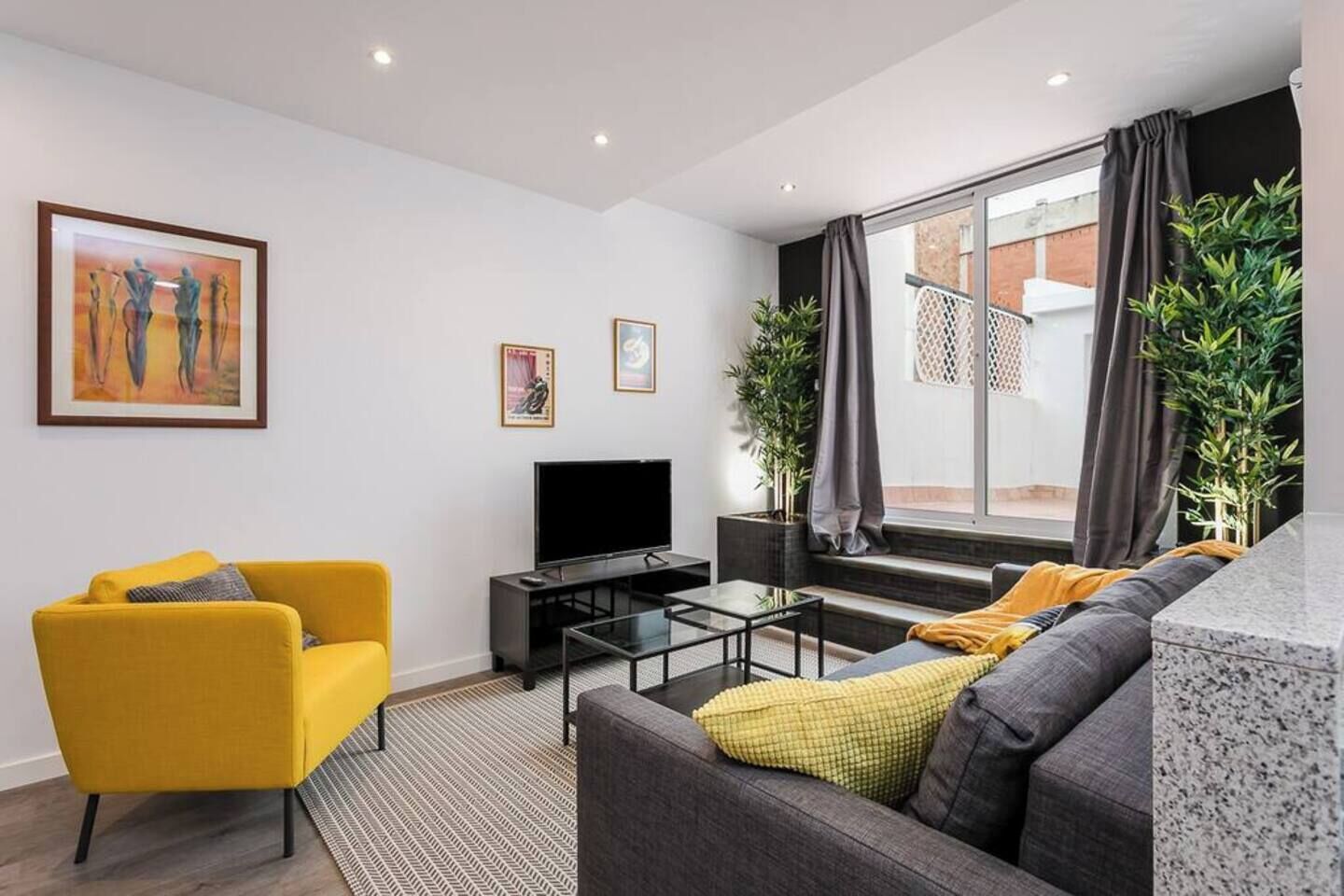 Un salon moderne avec un fauteuil jaune, un canapé gris, un téléviseur à écran plat et une grande fenêtre avec des rideaux.