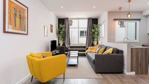 Ein modernes Wohnzimmer mit einer Couch, einem gelben Sessel und einem Couchtisch. Ein großes Fenster mit Vorhängen, eine Pflanze und ein Bild an der Wand sind ebenfalls zu sehen.