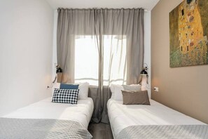Une chambre d’hôtel avec deux lits, une grande fenêtre avec des rideaux légers, deux lampes fixées au mur et un tableau accroché au mur.