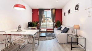 Een moderne woonkamer met een witte bank, een glazen salontafel, een eettafel met stoelen en een rode hanglamp.
