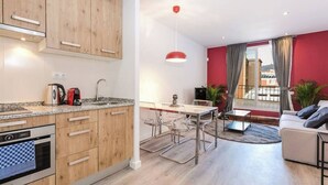 Een moderne keuken met houten kastjes en een inbouwoven, een eethoek met een tafel en stoelen, en een woonkamer met een bank en uitzicht op de stad door grote ramen.