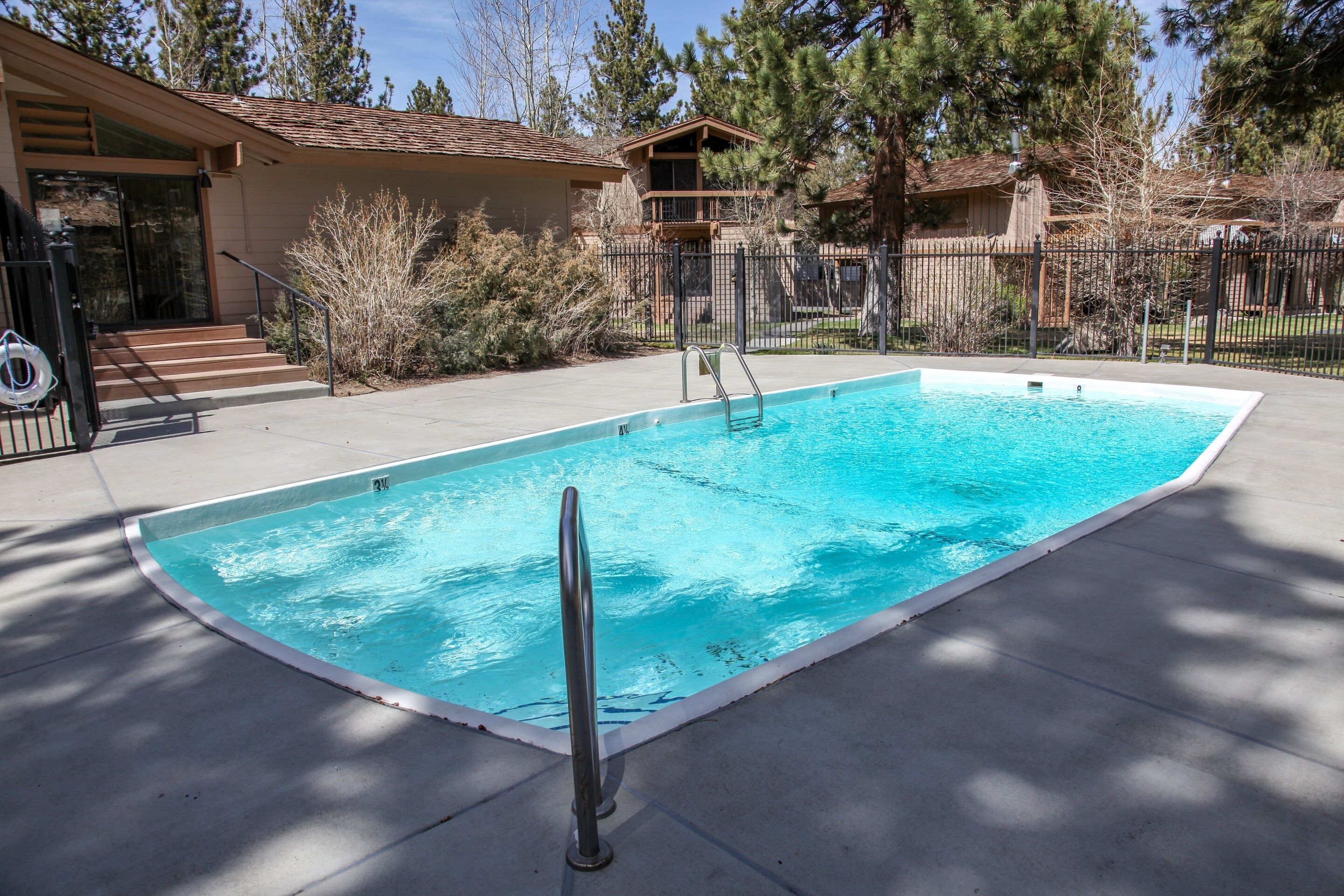 A rectangular swimming pool with a metal ladder, surrounded by a concrete deck and a metal fence.