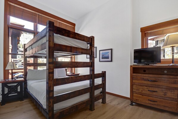 A bunk bed with a wooden frame, a bedside table with a lamp, a dresser with drawers, and a television on top.