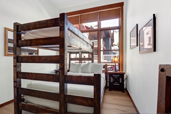 Una cama litera con estructura de madera y ropa de cama blanca, una mesita con lámpara, y una chimenea en una habitación con ventanas amplias y cuadros enmarcados en la pared.