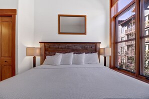 A neatly made bed with a wooden headboard, two bedside lamps, and a large window offering a view of buildings outside.