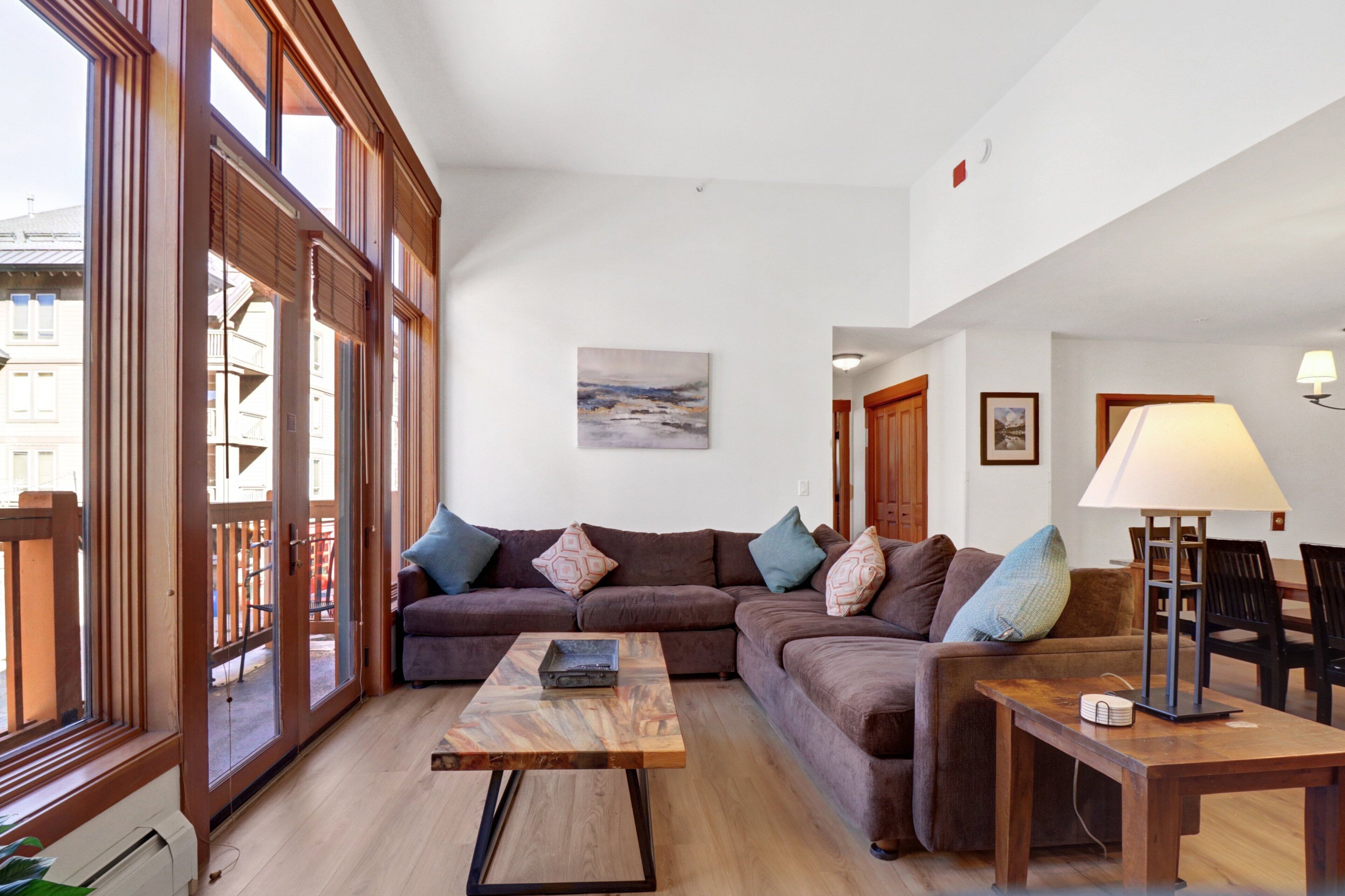A spacious living room with a large window, a wooden coffee table, a brown sofa, and a lamp.