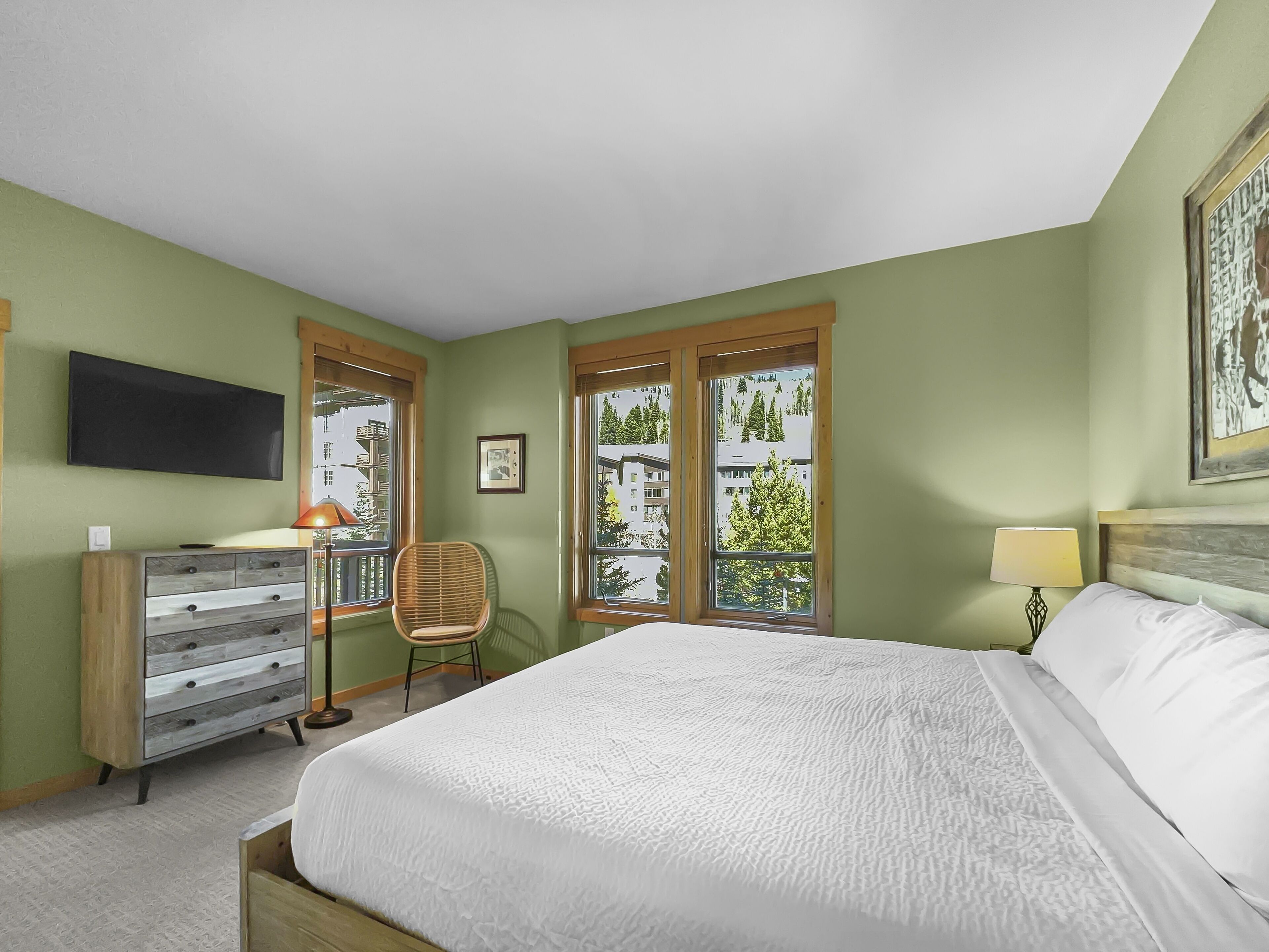 A bedroom with a bed, a dresser, a chair, a TV, and windows with a view of trees and buildings.