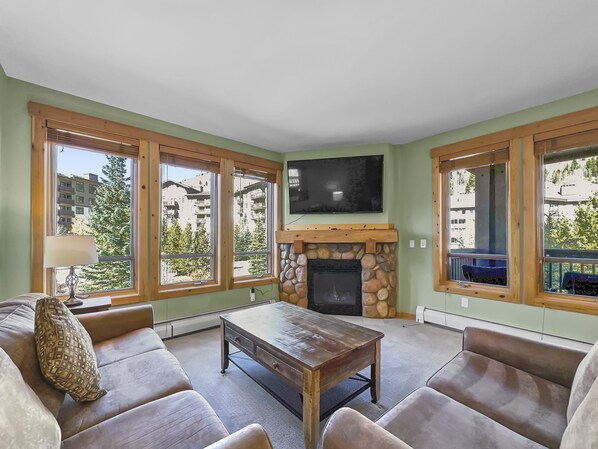 A living room with a stone fireplace, a television mounted above it, and large windows offering a view of buildings and trees. - Tx205 Taylors Crossing 2 Bedroom Condo by RedAwning (Copper Mountain)