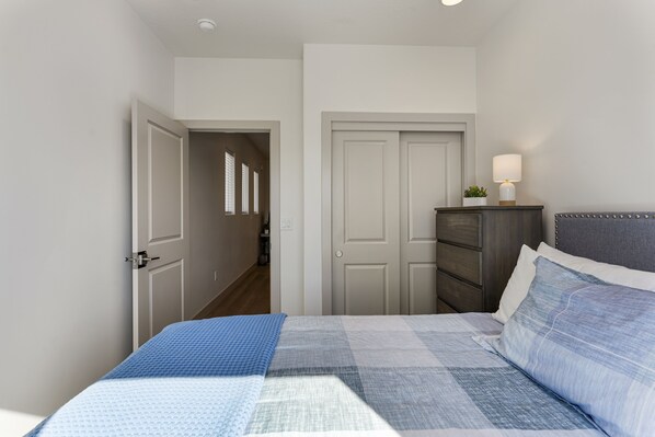 A bedroom with a bed, a dresser, a lamp, and a door leading to another room. - Hallser's Hive #4 3 Bedroom Townhouse (Santa Clara)