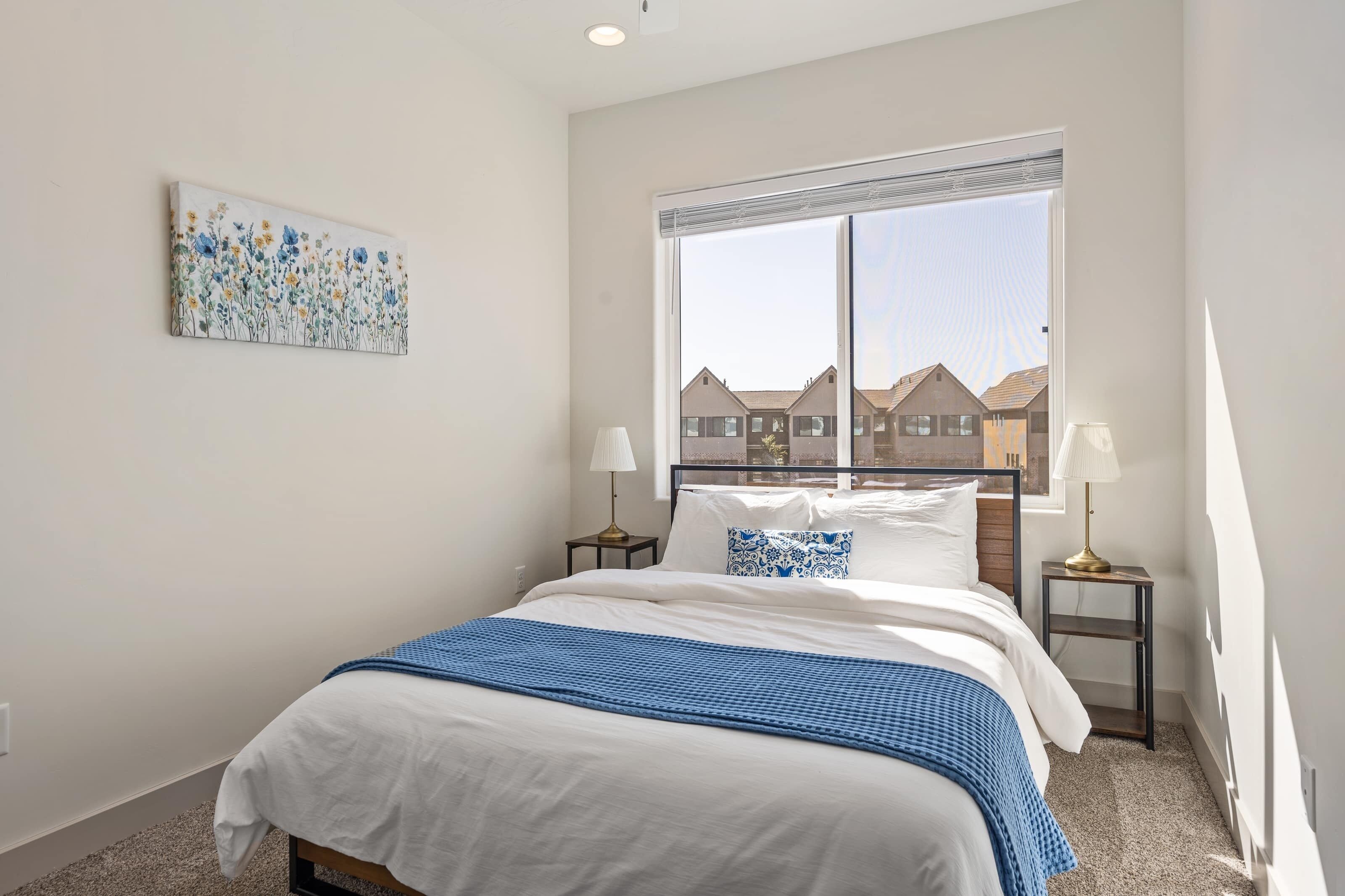 A bedroom with a large window, a bed with white and blue bedding, two bedside tables with lamps, and a floral artwork on the wall.