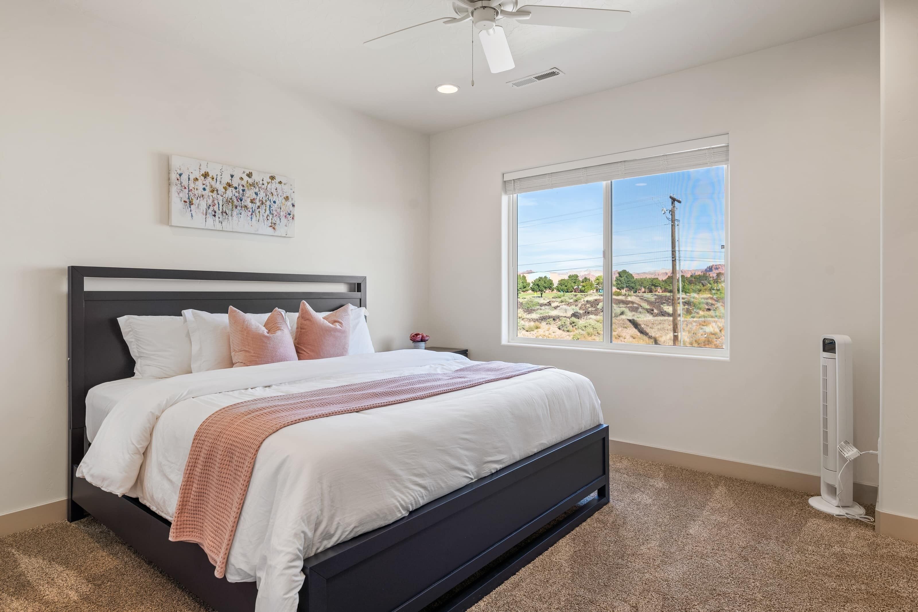 A bedroom with a bed, a ceiling fan, a window with a view, and a wall-mounted heater.