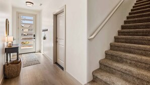Ein Flur mit Holzboden, einer mit Teppich belegten Treppe, einer Tür mit Fensterteil und einem kleinen Tisch mit Lampe und Korb.