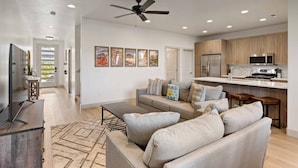 A modern living room with a sofa, coffee table, and a TV. The room has a ceiling fan, recessed lighting, and a kitchen area with wooden cabinets and a microwave. - Hallser's Escape 6 Bedroom Home (Santa Clara)