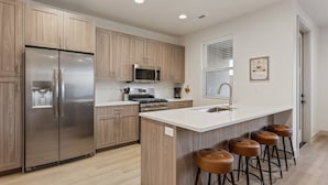 A modern kitchen with wooden cabinets, stainless steel appliances, a central island with stools, and a window with blinds. - Hallser's Escape 6 Bedroom Home (Santa Clara)