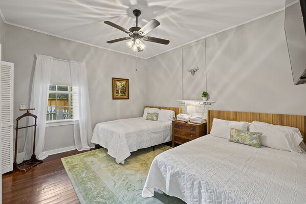 Un dormitorio con dos camas, ventilador de techo, ventana con cortinas, mesita de noche con lámpara y cabecero de madera.