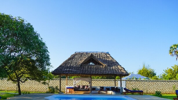 A swimming pool with a thatched-roof structure, lounge chairs, and a stone wall. - Villa m. Pool, Tropischen Garten & ca 7 Minuten zum Herrlichen Weißen Sandstrand (Msambweni)