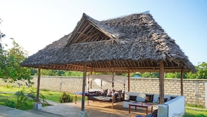 A covered outdoor seating area with a thatched roof, stone walls, and a stone floor. - Villa m. Pool, Tropischen Garten & ca 7 Minuten zum Herrlichen Weißen Sandstrand (Msambweni)