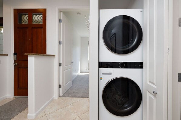 A laundry room with two stacked front-loading washing machines, a wooden door, and a hallway leading to another room.