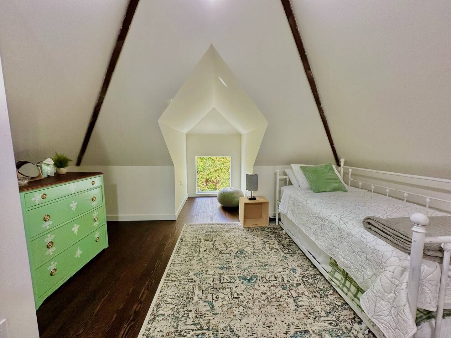 A bedroom with a sloped ceiling, a bed with a white headboard, a green dresser, and a patterned rug.