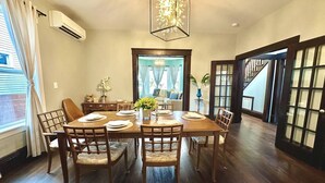 Une salle à manger avec une table en bois dressée pour six personnes, entourée de chaises. Un lustre est suspendu au-dessus de la table, et une fenêtre avec des rideaux se trouve à gauche.