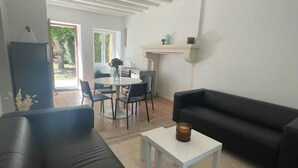 A living room with a black sofa, a white coffee table, and a black armchair. There is a dining area with a table and chairs, and a fireplace. - Charmante Maison à la Campagne (Sainte-Fauste)