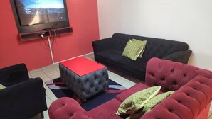 A living room with a flat-screen TV mounted on the wall, a red ottoman, a black sofa, and a red couch.