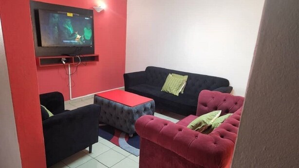 A living room with a flat-screen TV mounted on the wall, a red accent wall, and three sofas with green cushions.