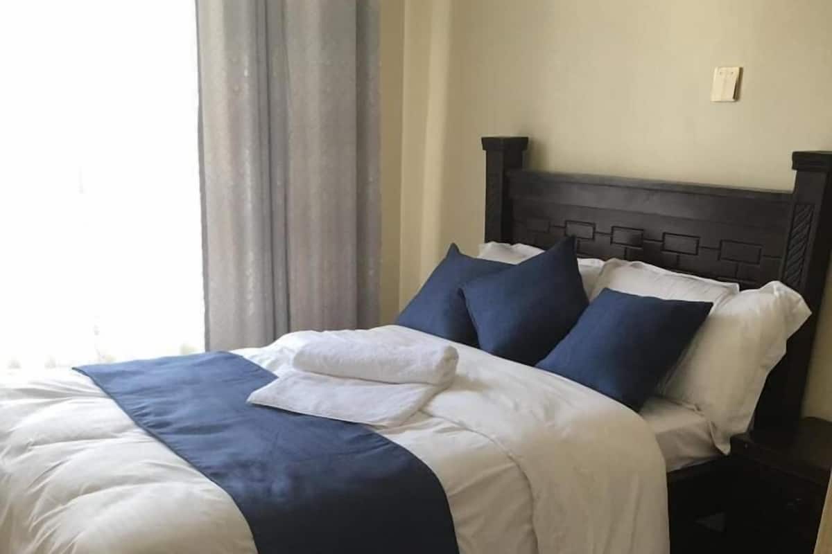 Una cama bien hecha con ropa de cama blanca y azul, cabecera oscura y una alfombra mullida blanca sobre piso de madera.