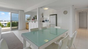 A modern kitchen with white cabinets, a glass dining table, and a view of the sea.